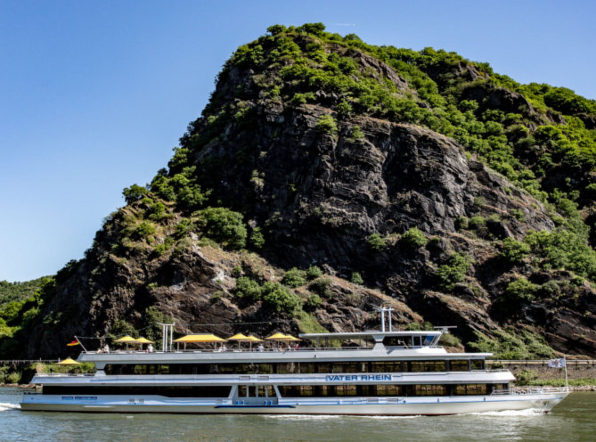 Das Schiff Vater Rhein vor dem Loreley-Felsen Auf dem Bild ist das weiße Ausflugsschiff „Vater Rhein“ zu sehen, das auf dem Rhein entlangfährt. Auf dem oberen Deck gibt es gelbe Sonnenschirme. Einige Menschen sitzen dort und schauen sich die Landschaft an. Im Hintergrund ist ein großer Felsen – der Loreley-Felsen. Der Himmel ist blau und die Sonne scheint.