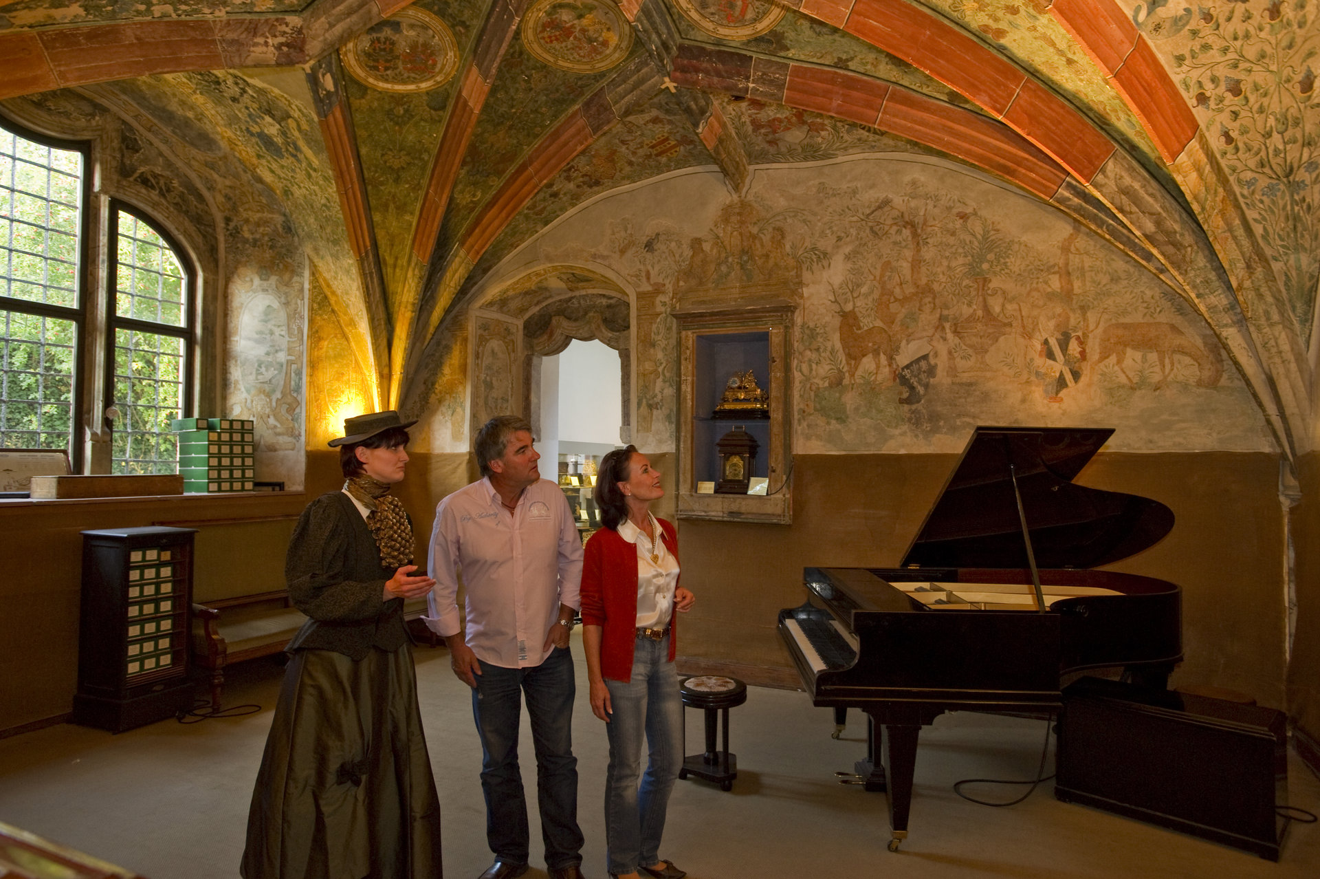 Musikkabinett Ein Paar wird von einer Dame im Gewand durch das Museum geführt. In einem schön ausgeleuchteten Raum steht ein Klavier.