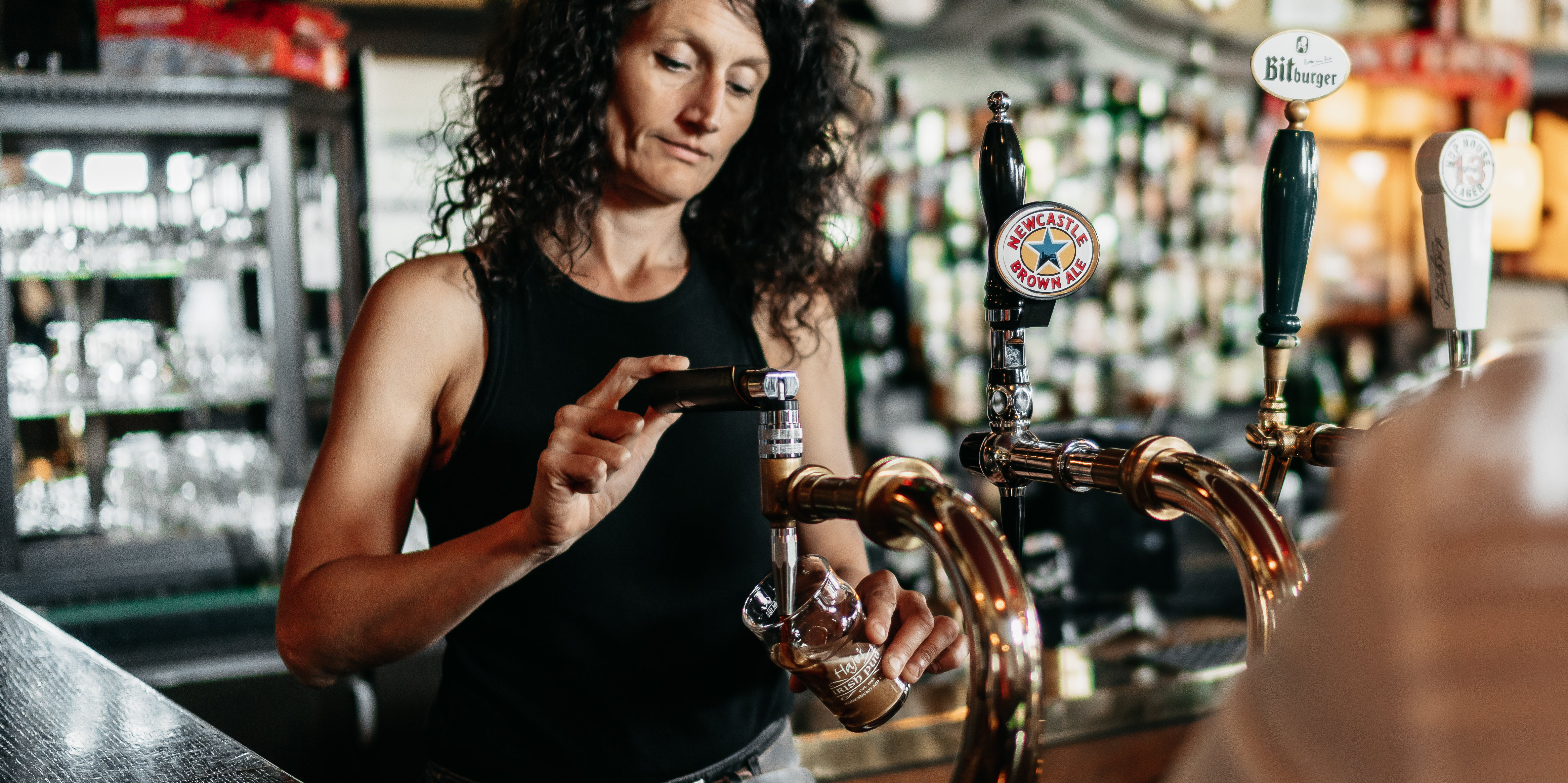 Eine Dame zapft ein dunkeles Bier hinterm Tresen, an dem ein Mann mit dem Rücken zur Kamera sitzt.