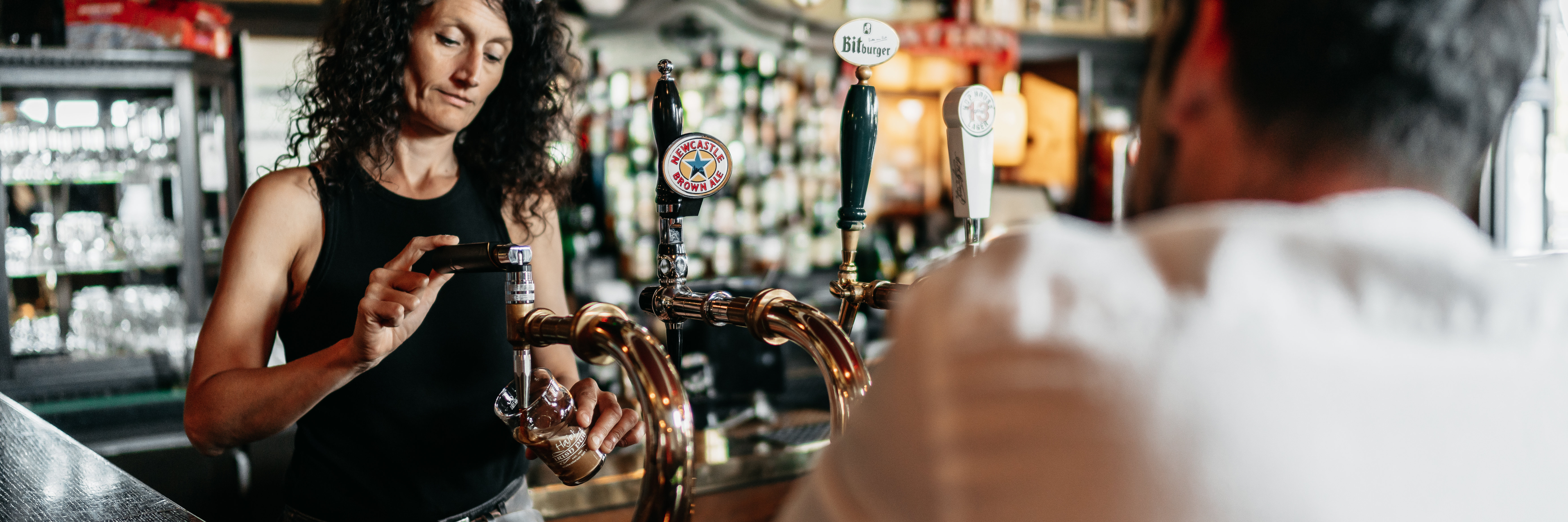 Eine Dame zapft ein dunkeles Bier hinterm Tresen, an dem ein Mann mit dem Rücken zur Kamera sitzt.