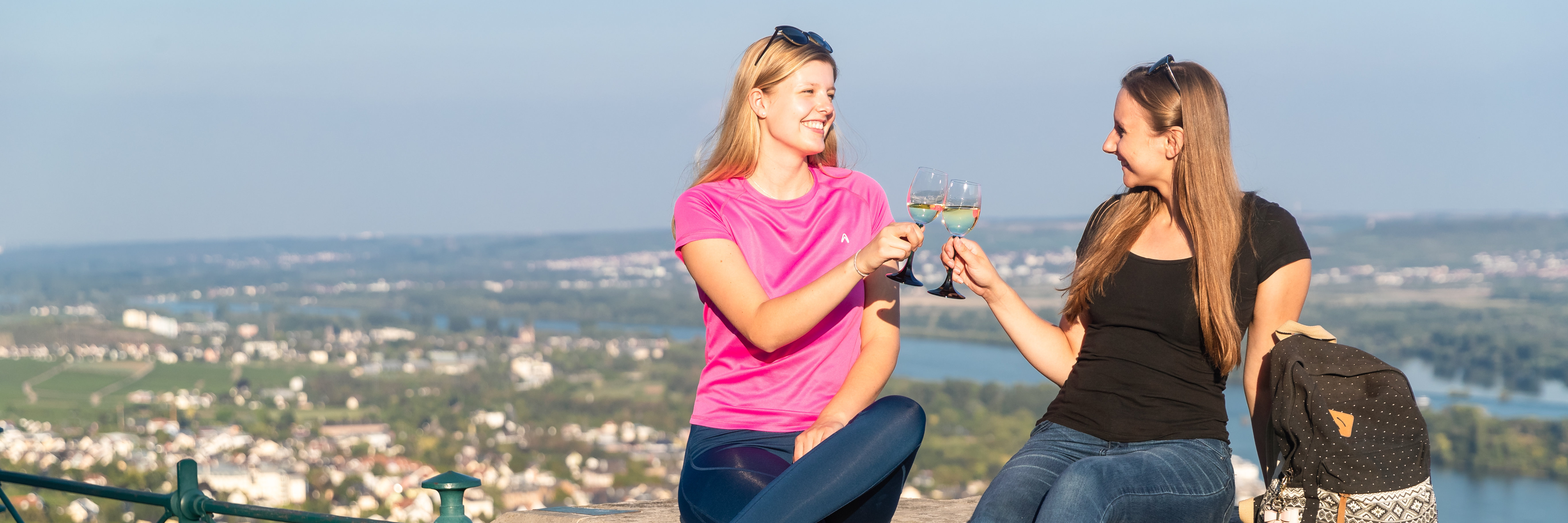 Weinwanderung Zwei junge Damen sitzen in sportlichen Outfits auf einer Mauer hoch über Rüdesheim und stoßen mit einem Glas Weißwein an.