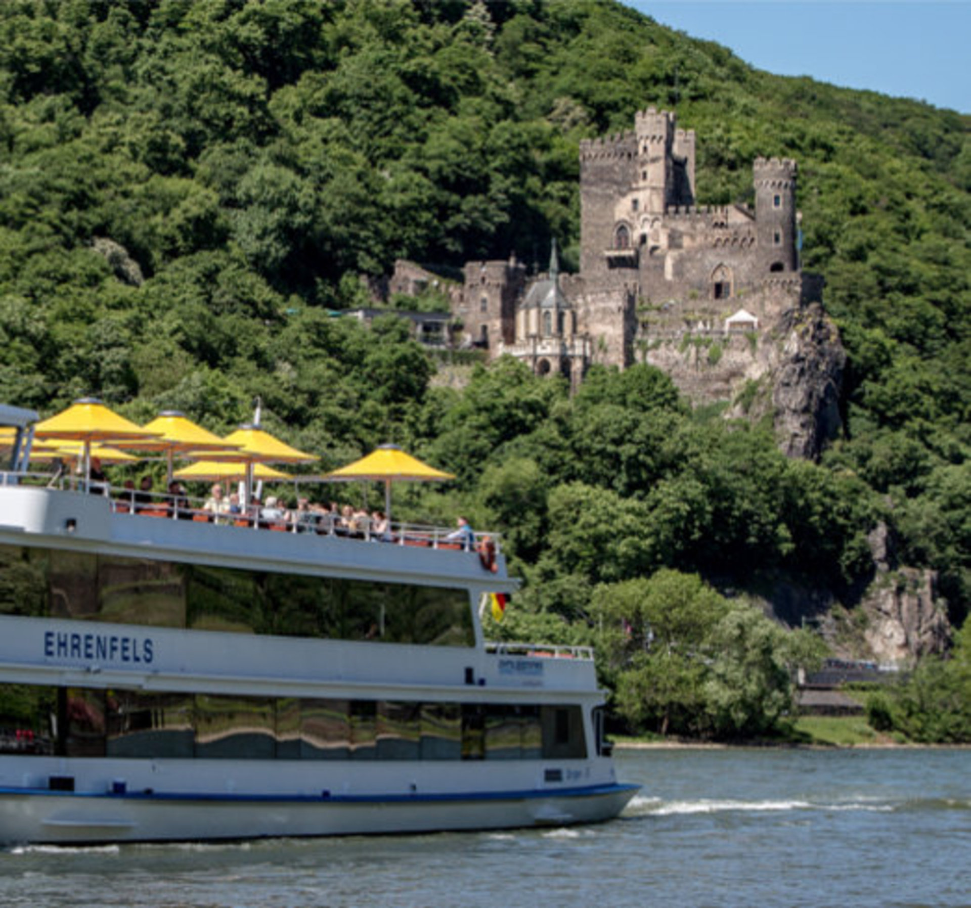 Schiff Ehrenfels Das Schiff Ehrenfels fährt bei schönem Wetter mit Gästen auf dem Rhein entlang. Im Hintegrund sieht man die Burg Rheinstein.