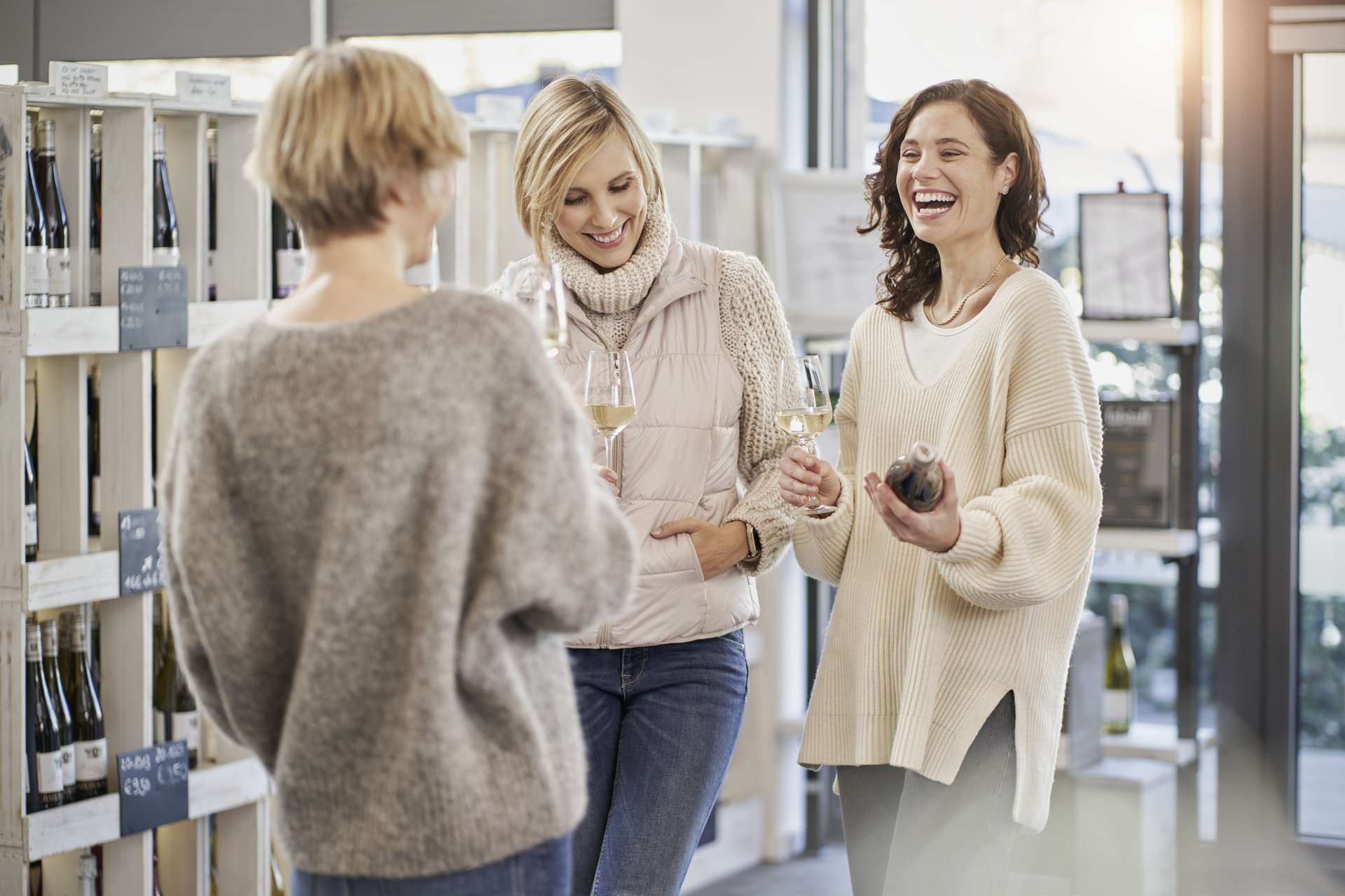 Weinausschank in Vinothek Drei Frauen stehen in einer Vinothek, halten Weingläser und lachen. Im Hintergrund sind Weinflaschen in Regalen zu sehen.