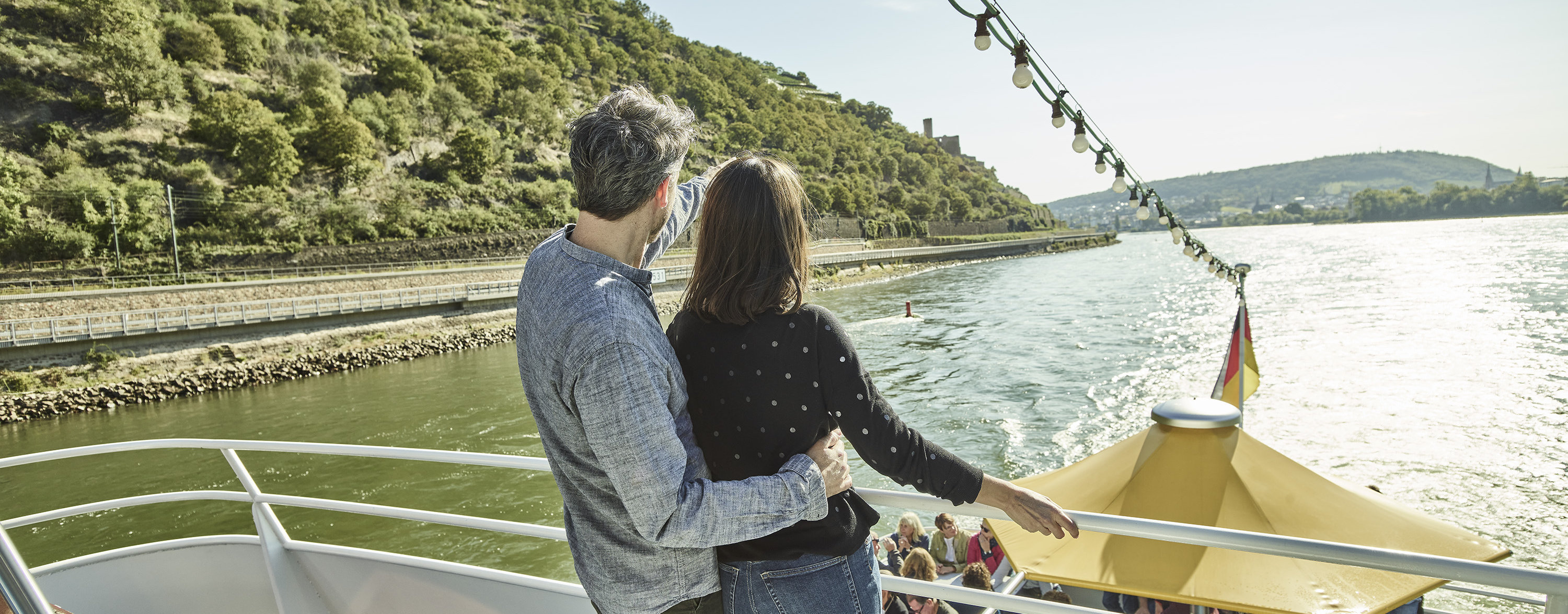 Schifffahrt auf dem Rhein Ein Paar steht bei Sonnenschein auf einem Schiff und blickt auf das Wasser und die Weinberge.