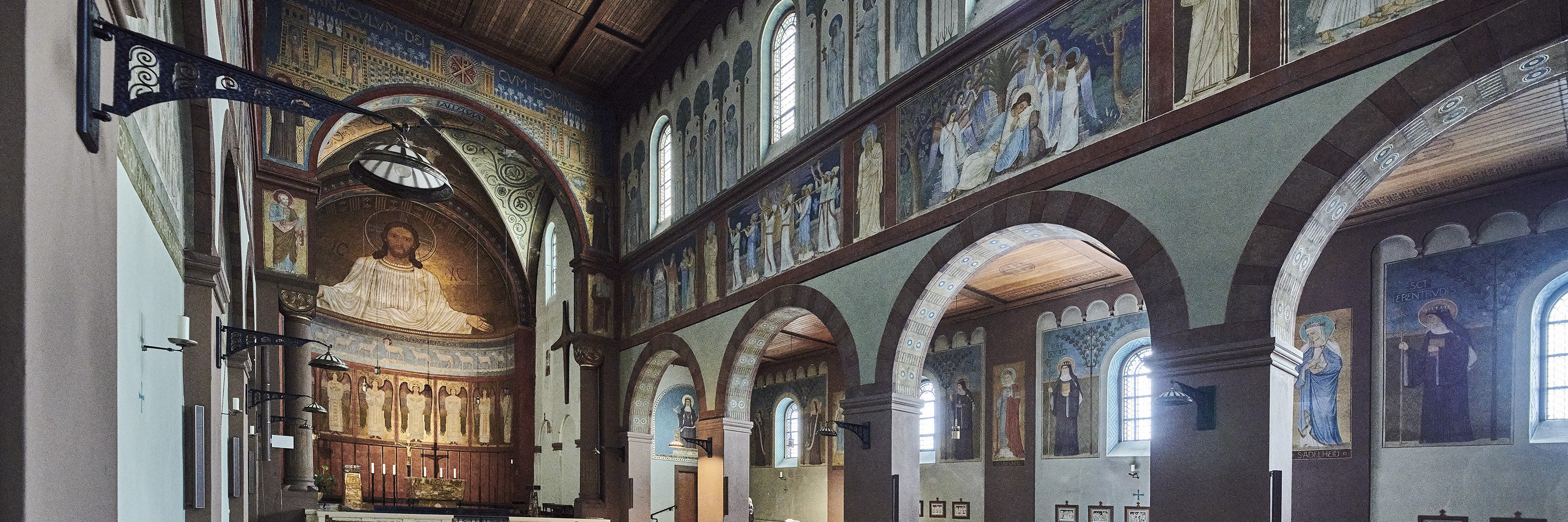 Abtei St. Hildegard Innenraum der Abteikirche St. Hildegard in Rüdesheim, Blick auf den Altarraum mit großer Christus-Darstellung an der Wand. Die Kirche ist mit Holzdecken, Wandmalereien, Rundbögen und Kirchenbänken gestaltet.