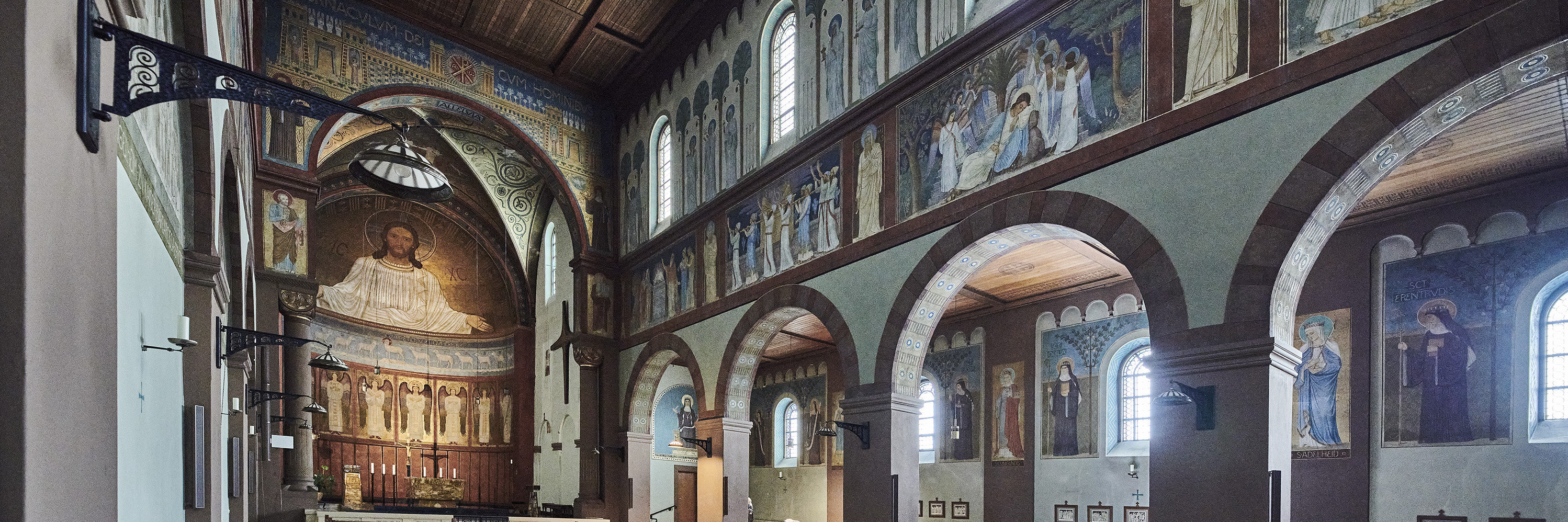 Abtei St. Hildegard  Innenraum der Abteikirche St. Hildegard in Rüdesheim, Blick auf den Altarraum mit großer Christus-Darstellung an der Wand. Die Kirche ist mit Holzdecken, Wandmalereien, Rundbögen und Kirchenbänken gestaltet.