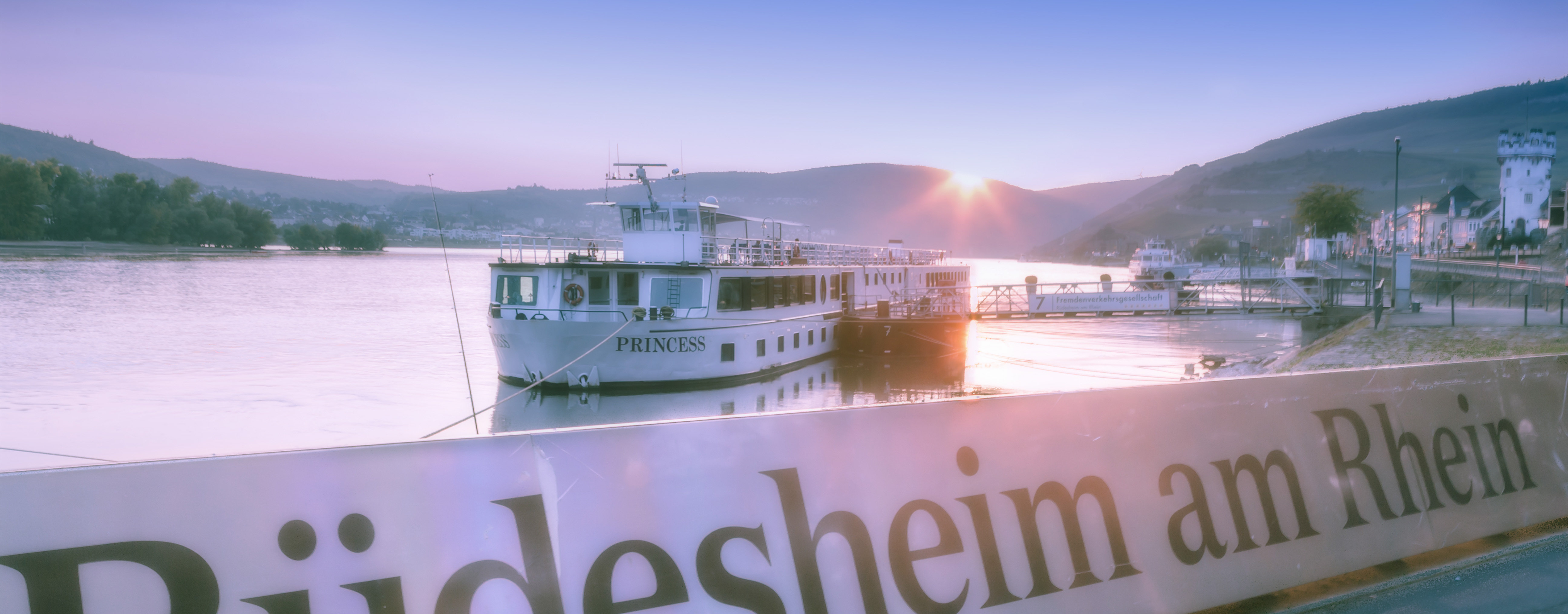 Rheinpromenade in Rüdesheim Das Bild zeigt die Uferpromenade von Rüdesheim am Rhein in der Abenddämmerung. Im Vordergrund ist ein Anlegesteg mit einem festgemachten Passagierschiff zu sehen, auf dem der Name „Princess“ steht.  Auf der rechten Seite erkennt man Teile der Altstadt und den Adlerturm.