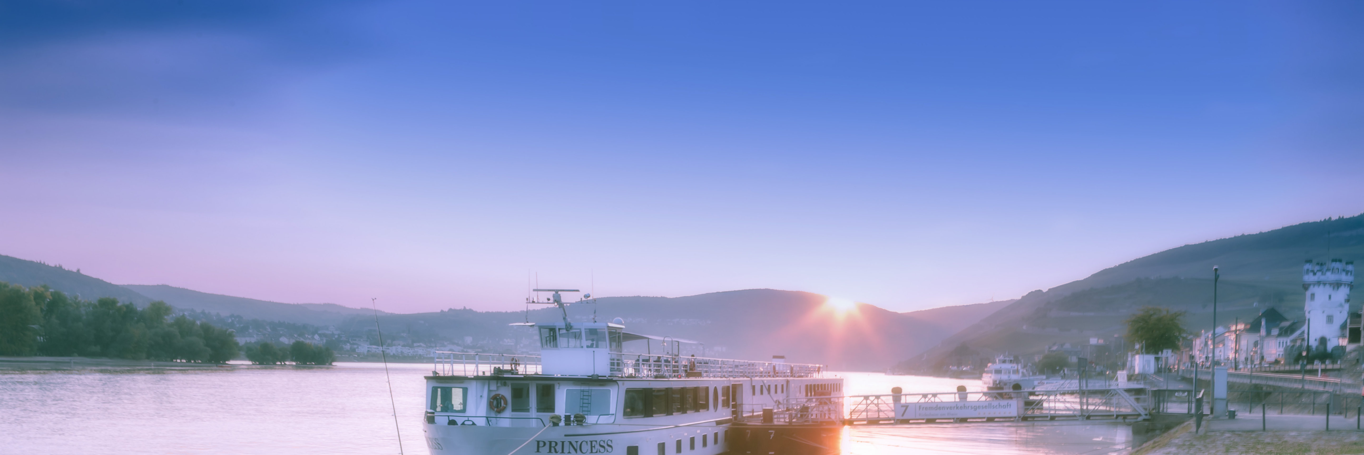 Rheinpromenade in Rüdesheim Das Bild zeigt die Uferpromenade von Rüdesheim am Rhein in der Abenddämmerung. Im Vordergrund ist ein Anlegesteg mit einem festgemachten Passagierschiff zu sehen, auf dem der Name „Princess“ steht. Auf der rechten Seite erkennt man Teile der Altstadt und den Adlerturm.