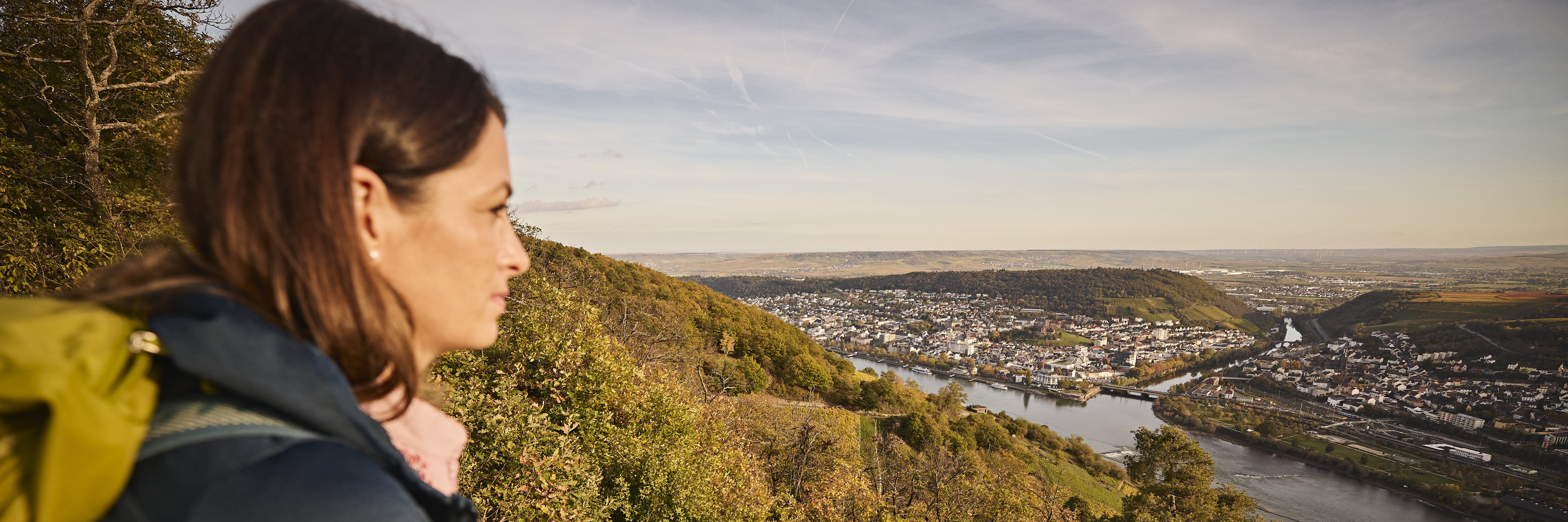 Aussicht über den Rhein Eine Frau lehnt an einer Steinmauer und blickt auf den Rhein, umgeben von grünen Hügeln, mit der Stadt Bingen am gegenüberliegenden Ufer bei sonnigem Himmel.