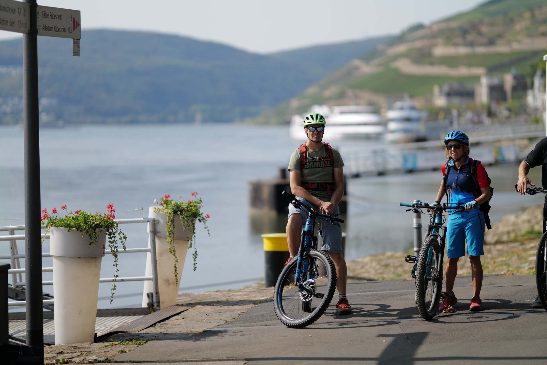 Fahrradfahren in Rüdesheim Ein Paar am Rhein macht eine Pause an der Rüdesheimer Rheinpromenade. Der Mann sitzt auf seinem Fahrradsitz und die Frau steht daneben und hält Ihr Fahrrad am Lenkrad fest. Im Hintergrund sieht man den Rhein, die Weinberge und zwei Schiffe.